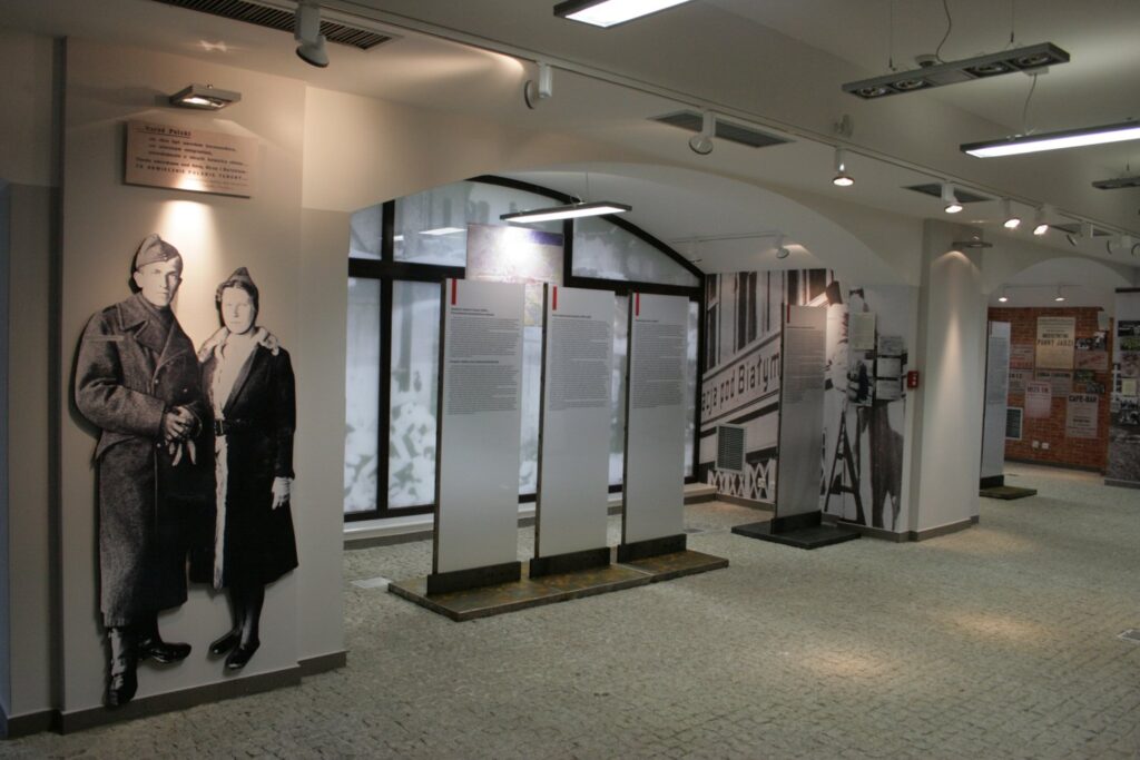 Widok sali ekspozycyjnej z planszami tematycznymi, fotografiami oraz elementami aranżacji historycznej wystawy (kliknięcie powiększy zdjęcie).