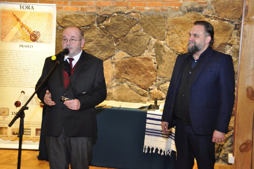 Dyrektor muzeum przemawia podczas otwarcia wystawy poświęconej historii Biblii, obok stoi kurator ekspozycji, a w tle widoczne są plansze edukacyjne oraz rekonstrukcja wnętrza zwojów Tory (kliknięcie powiększy zdjęcie).