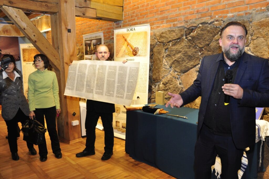Mężczyzna prezentuje uczestnikom jeden z najcenniejszych eksponatów – reprodukcję dawnego rękopisu Biblii – na tle plansz poświęconych Torze i historii świętych ksiąg (kliknięcie powiększy zdjęcie).