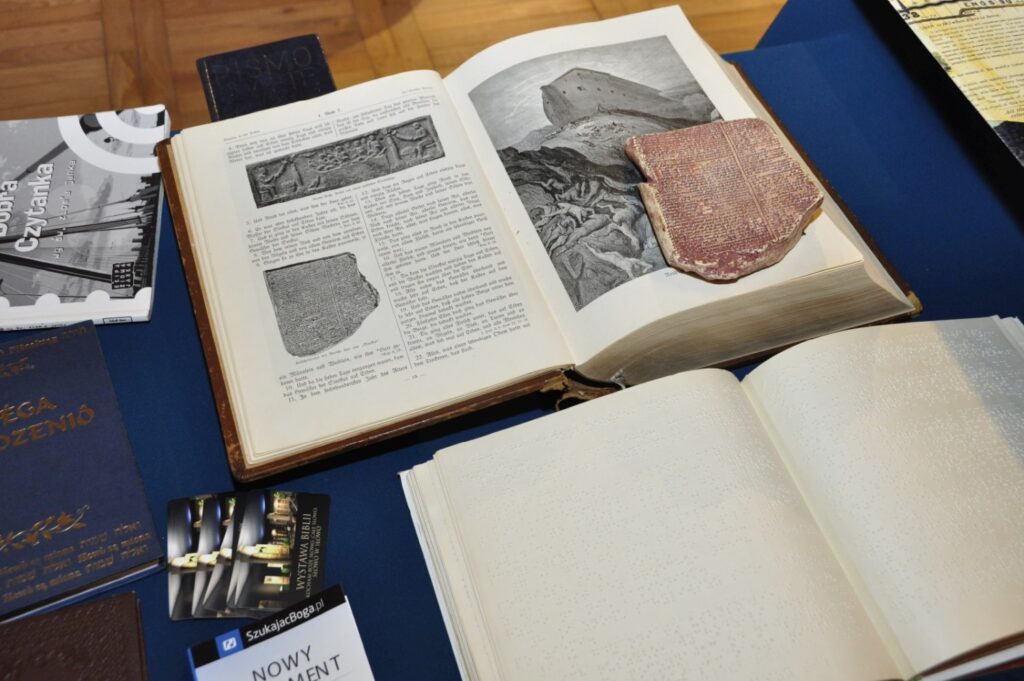 Zbliżenie na eksponaty prezentowane na wystawie – otwarta publikacja z ilustracjami, fragment glinianej tabliczki z pismem oraz współczesne wydanie Biblii zapisane alfabetem Braille’a (kliknięcie powiększy zdjęcie).