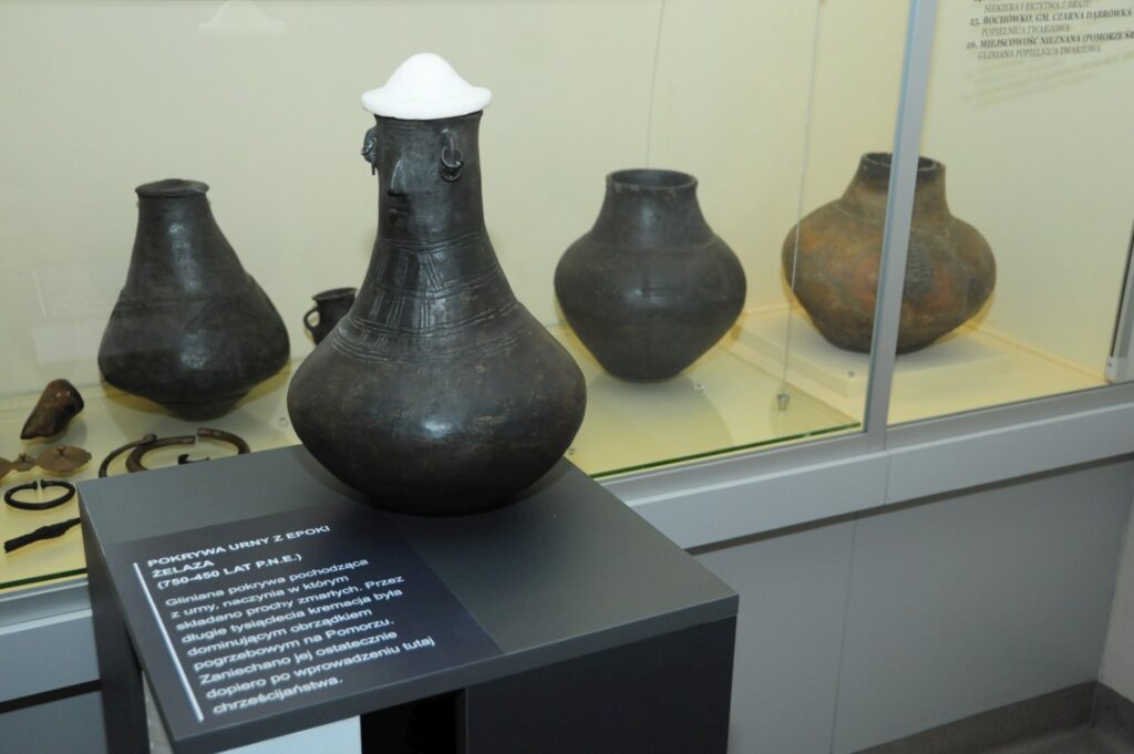 Zbliżenie na ceramiczne naczynie z epoki brązu prezentowane na postumencie, z gablotą pełną naczyń i innych zabytków archeologicznych widoczną w tle (kliknięcie powiększy zdjęcie).