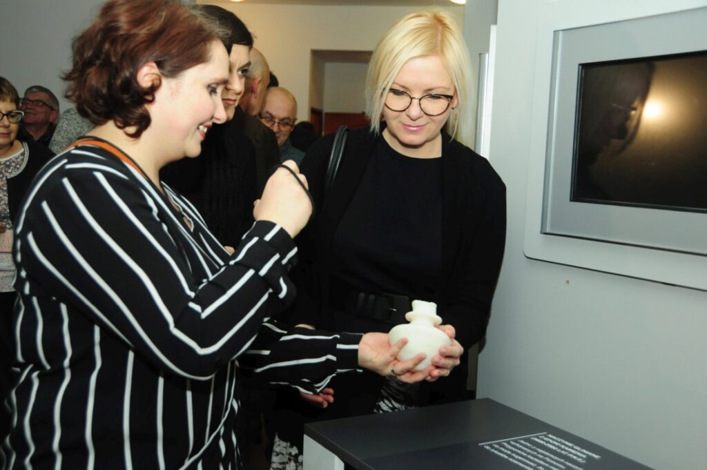Zwiedzające oglądają replikę niewielkiego glinianego kubka prezentowanego na wystawie, korzystając z przygotowanej lupy ułatwiającej dostrzeżenie detali przedmiotu (kliknięcie powiększy zdjęcie).