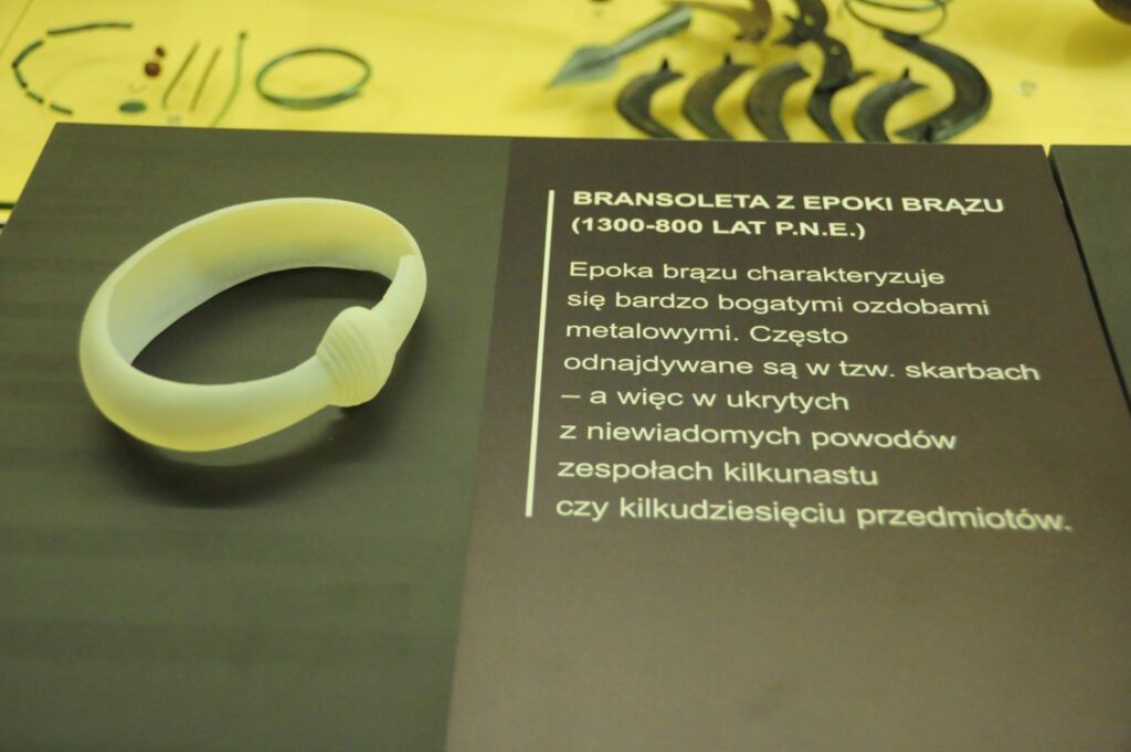 Zbliżenie na kopię bransolety z epoki brązu, wykonanej w druku 3D, prezentowaną wraz z opisem przedstawiającym jej datowanie, funkcję oraz okoliczności odkrywania podobnych ozdób podczas badań archeologicznych (kliknięcie powiększy zdjęcie).