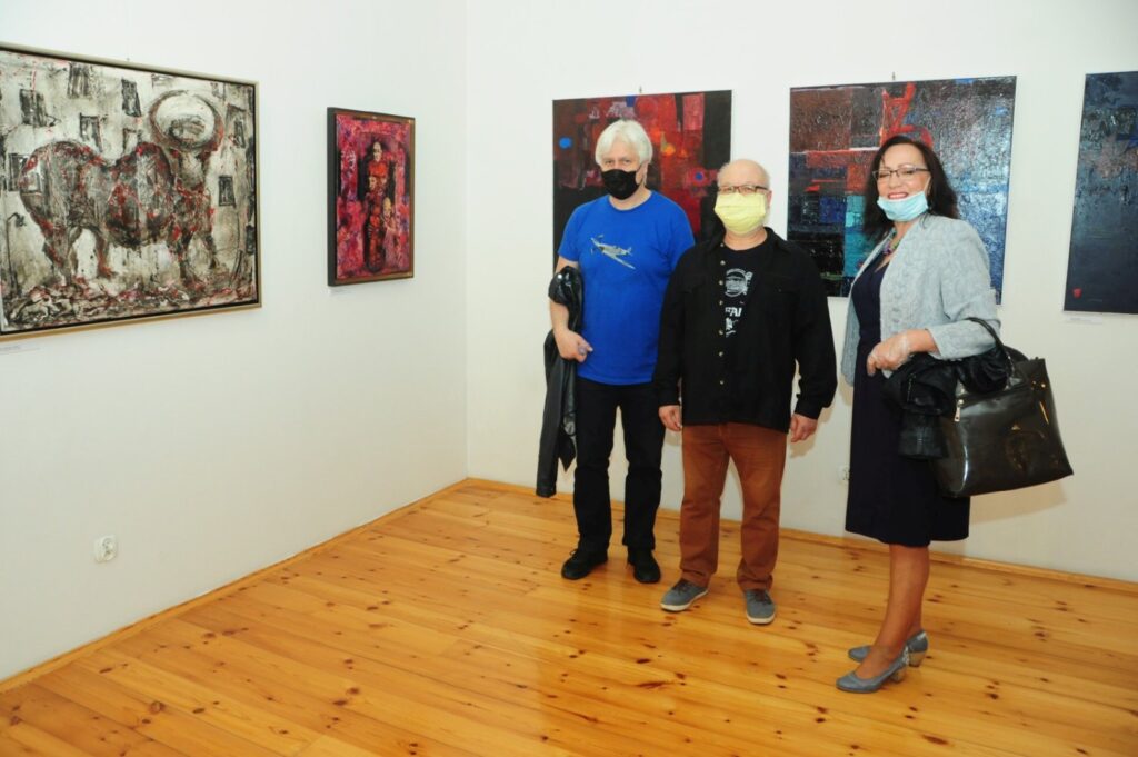 Trzy osoby stoją w sali wystawowej na tle dużych obrazów utrzymanych w ekspresyjnej, abstrakcyjnej stylistyce. Jasne wnętrze galerii z drewnianą podłogą i białymi ścianami podkreśla kontrastowe barwy prezentowanych prac (kliknięcie powiększy zdjęcie).