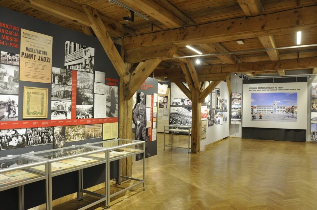 Szerokie ujęcie sali wystawowej poświęconej historii powojennego Koszalina. Na planszach prezentowane są archiwalne fotografie, dokumenty i teksty, a w gablotach umieszczono historyczne eksponaty związane z życiem miasta (kliknięcie powiększy zdjęcie).