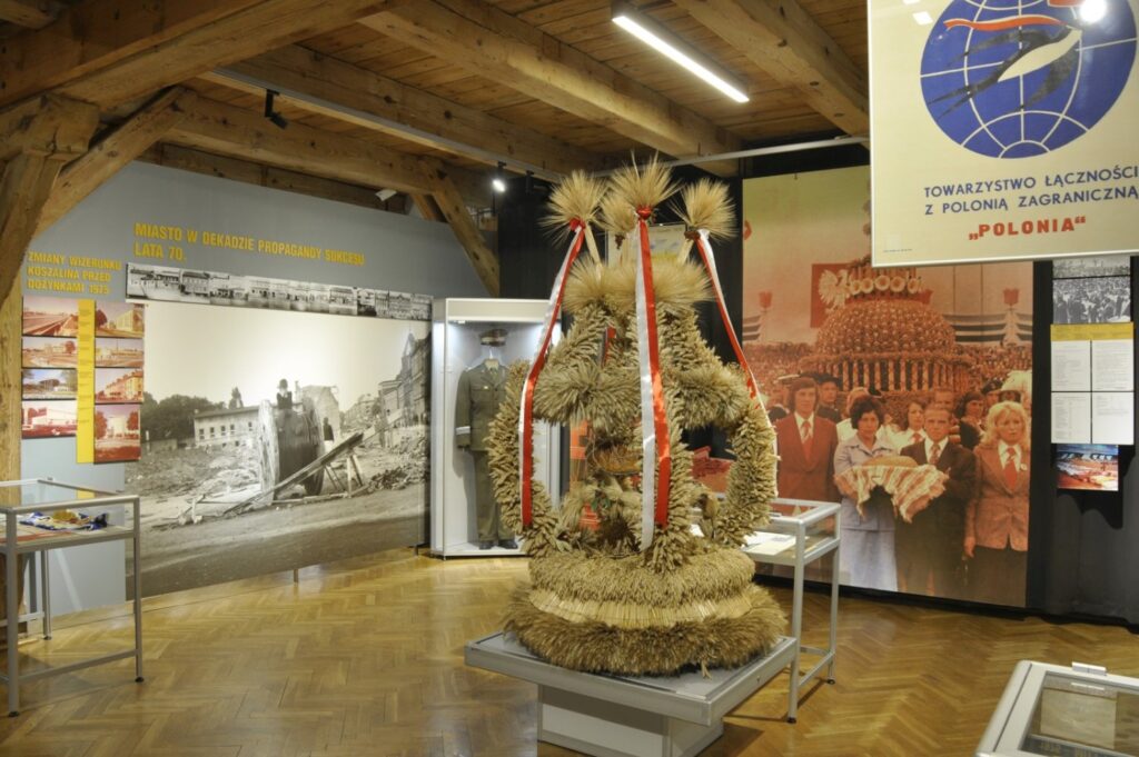Widok sali wystawowej z dużą tradycyjną palmą wielkanocną ustawioną na środku ekspozycji. W tle znajdują się plansze poświęcone historii miasta, fotografie mieszkańców oraz gabloty z eksponatami muzealnymi (kliknięcie powiększy zdjęcie).