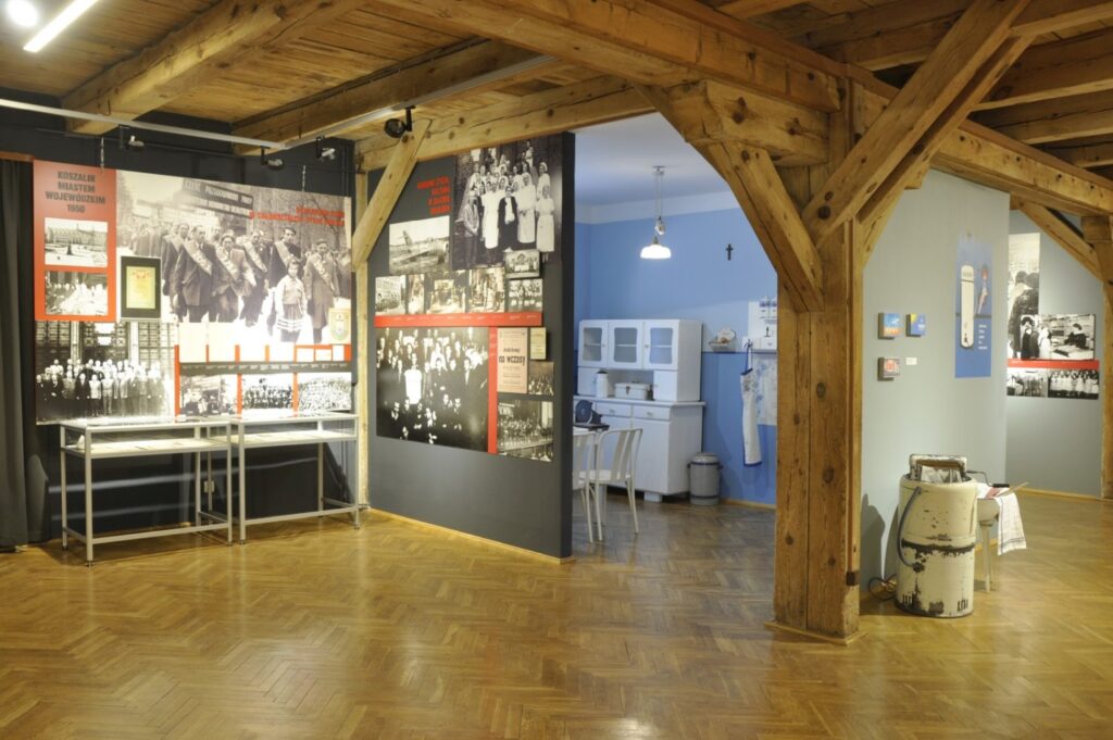 Szerokie ujęcie końcowej części wystawy historycznej. Drewniana konstrukcja wnętrza otacza plansze z archiwalnymi fotografiami, gabloty z dokumentami oraz zrekonstruowane wnętrze mieszkalne, ukazując różnorodność prezentowanych materiałów (kliknięcie powiększy zdjęcie).