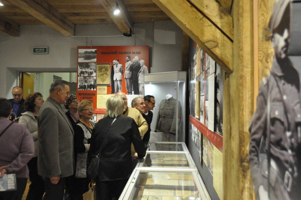 Zwiedzający oglądają fragment wystawy poświęcony historii powojennego miasta. Na ścianach znajdują się plansze z archiwalnymi fotografiami, a w gablotach prezentowane są dokumenty i pamiątki z epoki (kliknięcie powiększy zdjęcie).