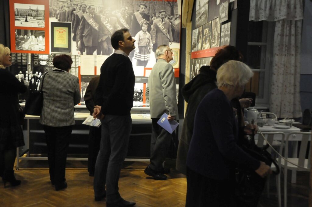 Goście zwiedzają końcową część ekspozycji historycznej, zatrzymując się przed planszami z archiwalnymi fotografiami i dokumentami. Wystawa prezentuje historię miasta poprzez fotografie, teksty oraz oryginalne pamiątki zgromadzone w gablotach (kliknięcie powiększy zdjęcie).