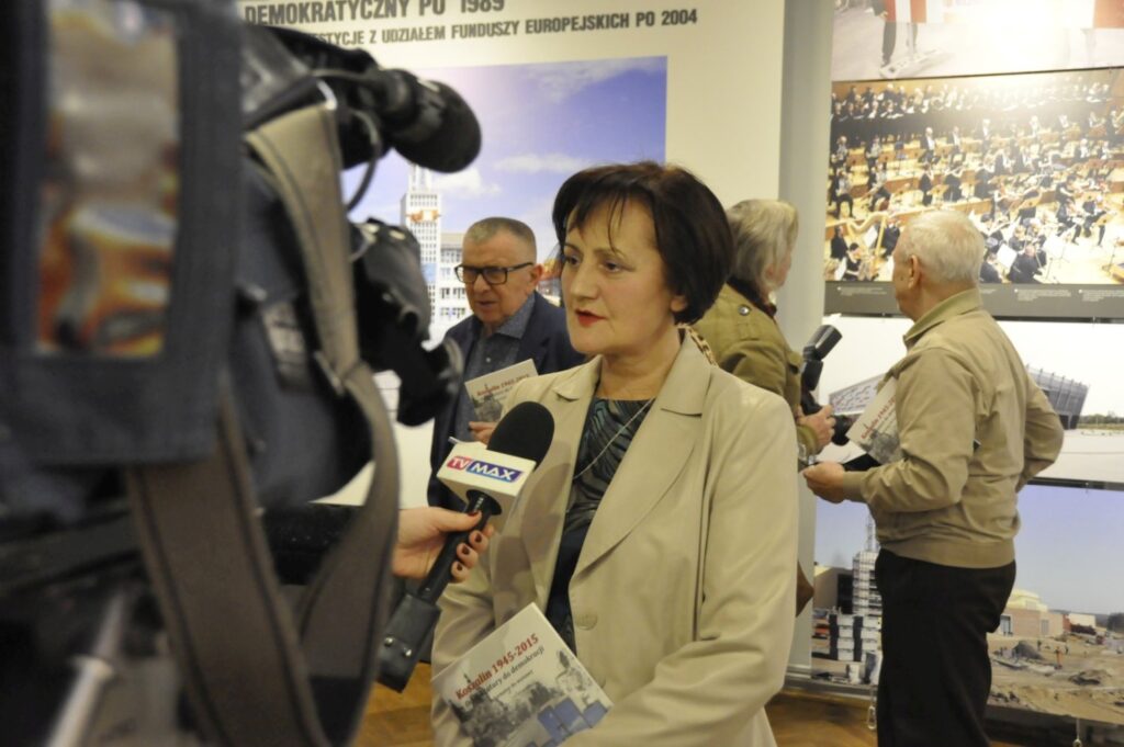 Wywiad przed kamerą telewizyjną realizowany podczas otwarcia wystawy. Dziennikarka rozmawia z kobietą na tle plansz prezentujących archiwalne fotografie i materiały dokumentujące historię miasta (kliknięcie powiększy zdjęcie).