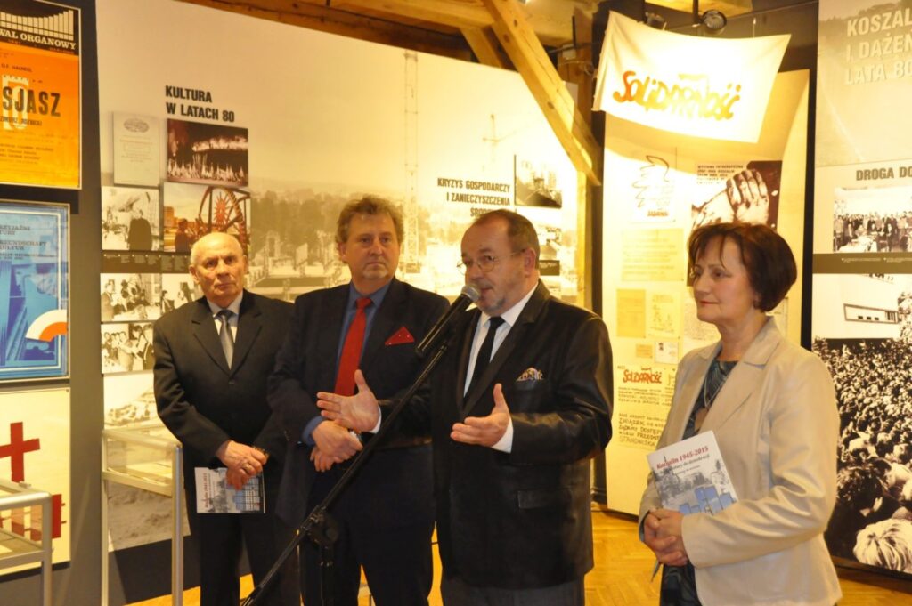 Oficjalne otwarcie wystawy. Przemawiający mężczyzna stoi przy mikrofonie w towarzystwie organizatorów i zaproszonych gości. W tle znajdują się plansze prezentujące fotografie, dokumenty i materiały poświęcone przemianom społecznym oraz historii miasta (kliknięcie powiększy zdjęcie).