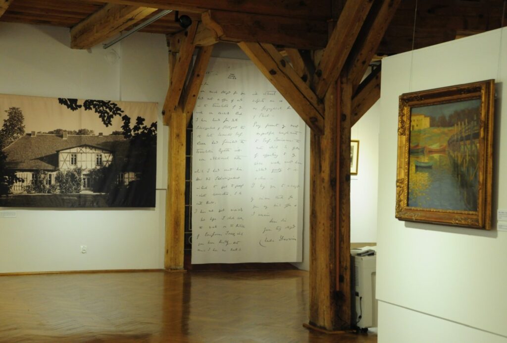 Fragment ekspozycji łączącej malarstwo z instalacją artystyczną. Obok obrazu przedstawiającego pejzaż znajduje się wielkoformatowa fotografia dawnego budynku oraz pionowa kompozycja z odręcznie zapisanym tekstem na białym tle (kliknięcie powiększy zdjęcie).
