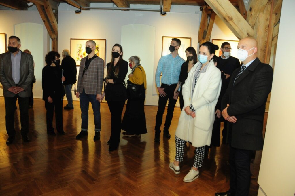 Goście wernisażu stoją w sali wystawowej, słuchając wystąpienia prowadzących. Na ścianach galerii eksponowane są obrazy w ramach, a przestrzeń wyznaczają drewniane filary historycznego budynku (kliknięcie powiększy zdjęcie).
