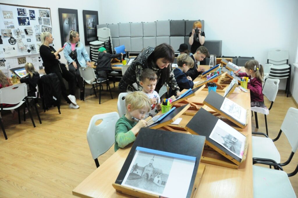 Sala warsztatowa z dziećmi pracującymi przy stołach nad zadaniami plastycznymi pod opieką dorosłych (kliknięcie powiększy zdjęcie).