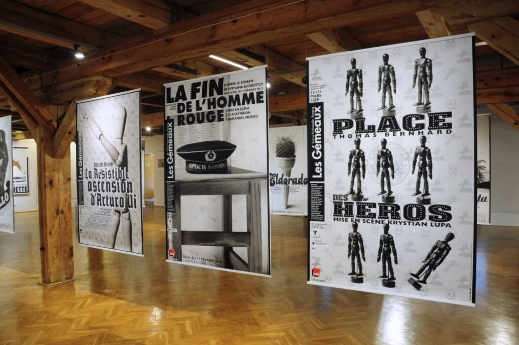 Fragment wystawy z plakatami „La fin de l'homme rouge” oraz „The Place of Heroes”; prace wykorzystują kontrast czerni i bieli oraz charakterystyczną typografię, a pomiędzy nimi umieszczono mniejsze elementy ekspozycji (kliknięcie powiększy zdjęcie).