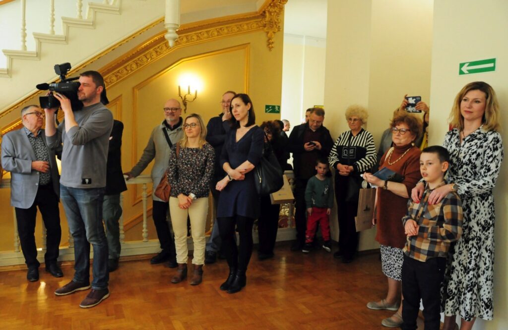 Grupa gości podczas wernisażu zgromadzona w holu muzeum; uczestnicy pozują do fotografii przy reprezentacyjnych schodach, wśród nich znajdują się osoby dorosłe i dzieci (kliknięcie powiększy zdjęcie).