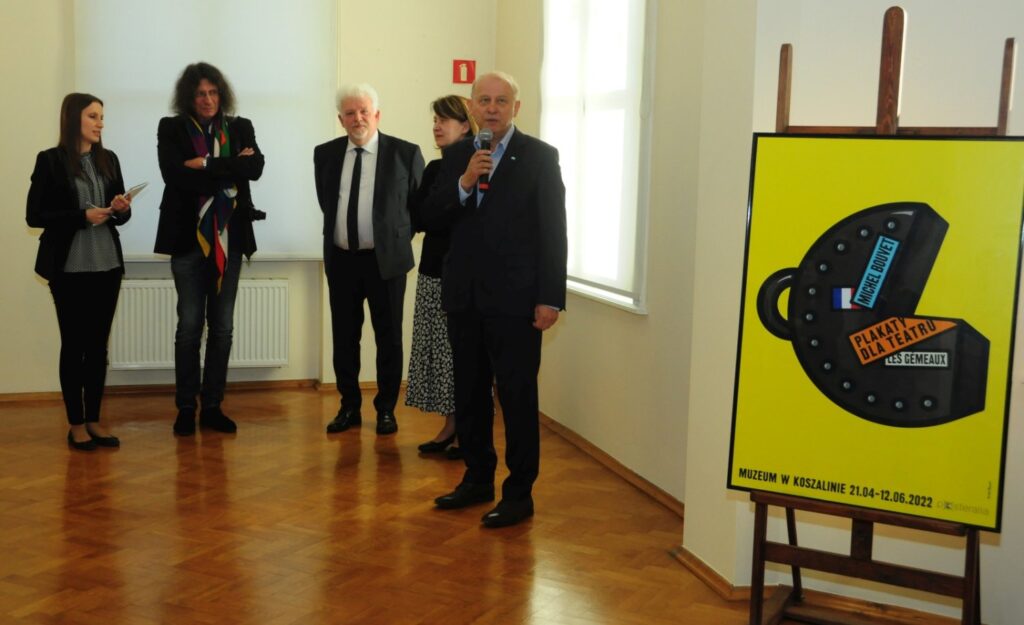 Fotografia z wernisażu przedstawiająca moment oficjalnego otwarcia wystawy; przy ścianie stoją uczestnicy wydarzenia, a na pierwszym planie widoczny jest plakat promujący ekspozycję (kliknięcie powiększy zdjęcie).