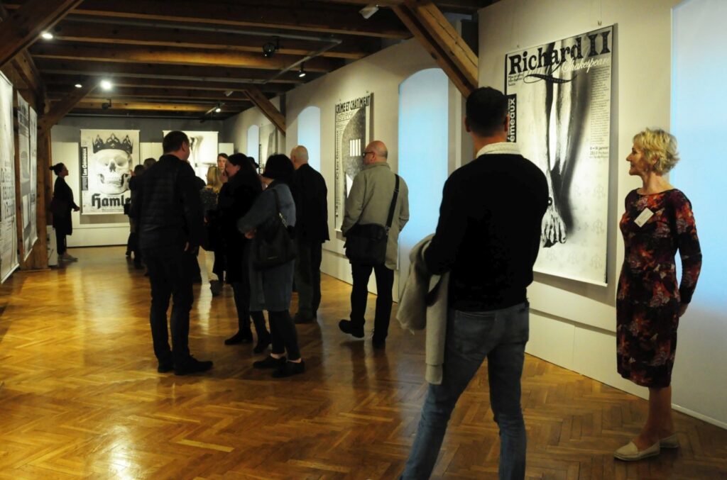 Zwiedzający podczas oglądania ekspozycji; w przestrzeni wystawowej widoczne są wielkoformatowe plakaty, w tym prace „Hamlet” i „Richard II”, a goście przemieszczają się pomiędzy prezentowanymi dziełami (kliknięcie powiększy zdjęcie).