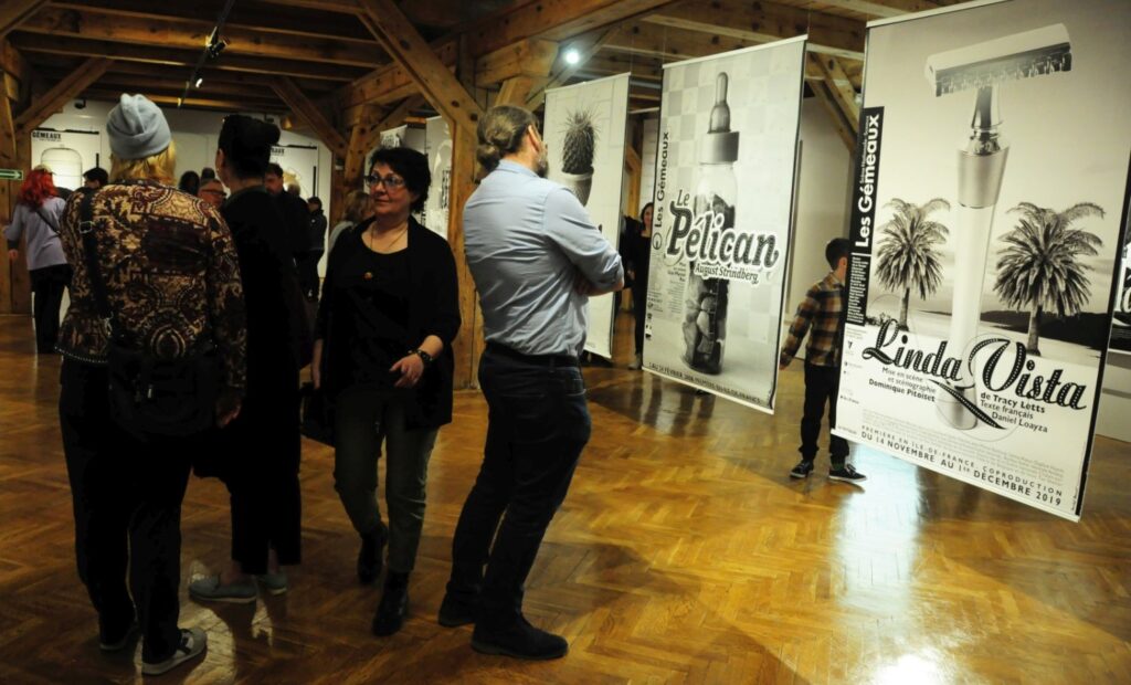 Zwiedzający podczas oglądania kolejnej części wystawy; w tle widoczne są plakaty „Pelican” oraz „Linda Vista”, a uczestnicy wernisażu zatrzymują się przy prezentowanych pracach (kliknięcie powiększy zdjęcie).