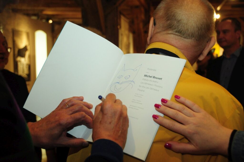 Zbliżenie na moment podpisywania publikacji podczas wernisażu; osoba trzyma otwartą książkę z odręczną dedykacją i rysunkiem, a druga osoba wpisuje dedykację na stronie (kliknięcie powiększy zdjęcie).