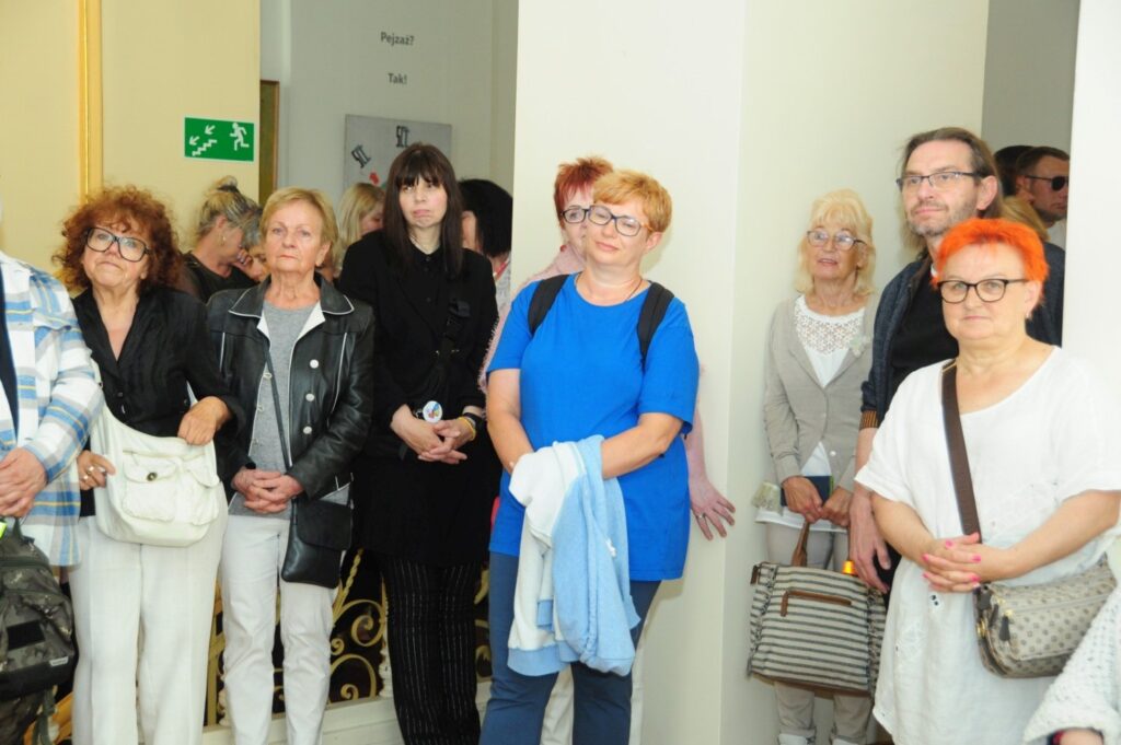 Grupa osób stoi razem w korytarzu galerii, pozując do zdjęcia; za nimi widoczne są drzwi, schody i fragmenty wystawy (kliknięcie powiększy zdjęcie).