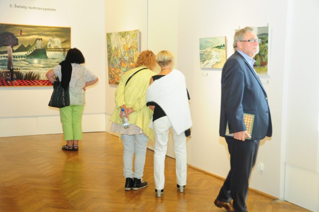 Zwiedzający oglądają obrazy na ścianach galerii; kilka osób stoi przed eksponatami, a po prawej stronie znajduje się mężczyzna trzymający katalog (kliknięcie powiększy zdjęcie).
