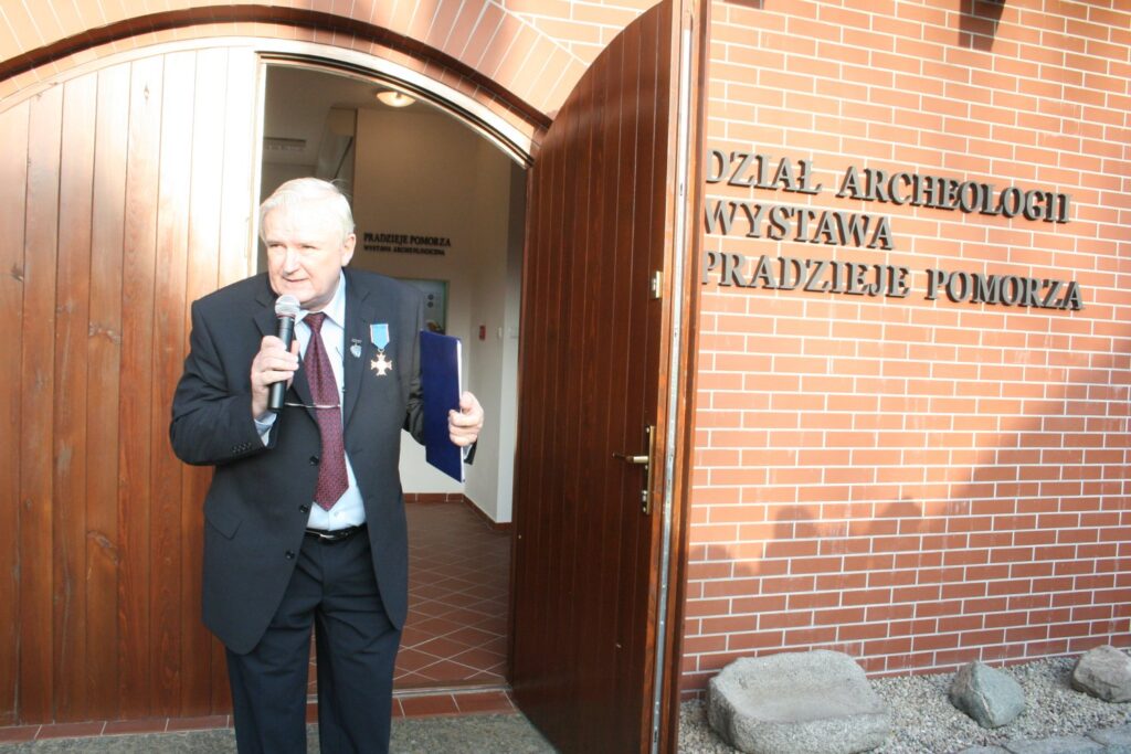 Mężczyzna z mikrofonem stoi przy wejściu do budynku z napisem Dział Archeologii Wystawa Pradzieje Pomorza i przemawia do zebranych (kliknięcie powiększy zdjęcie)