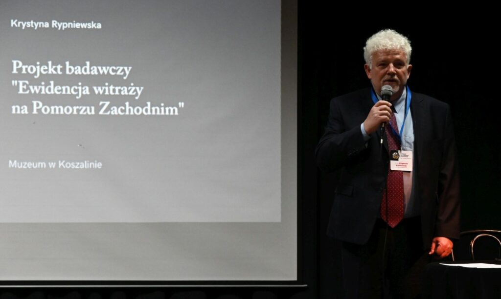 Mężczyzna przemawia do mikrofonu podczas konferencji naukowej, obok widoczny ekran z prezentacją (kliknięcie powiększy zdjęcie).
