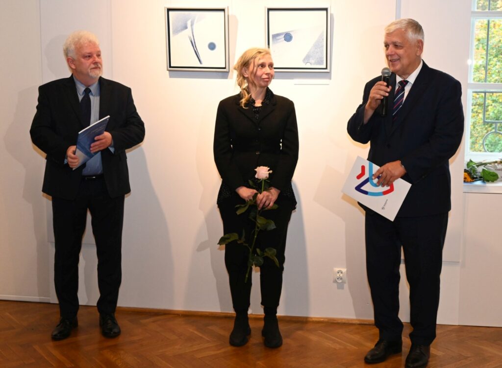 Trzy osoby stoją obok siebie; mężczyzna przemawia z mikrofonem, obok stoi kobieta z różą oraz drugi mężczyzna trzymający materiały związane z wystawą, a na ścianie w tle prezentowane są prace artystyczne (kliknięcie powiększy zdjęcie).