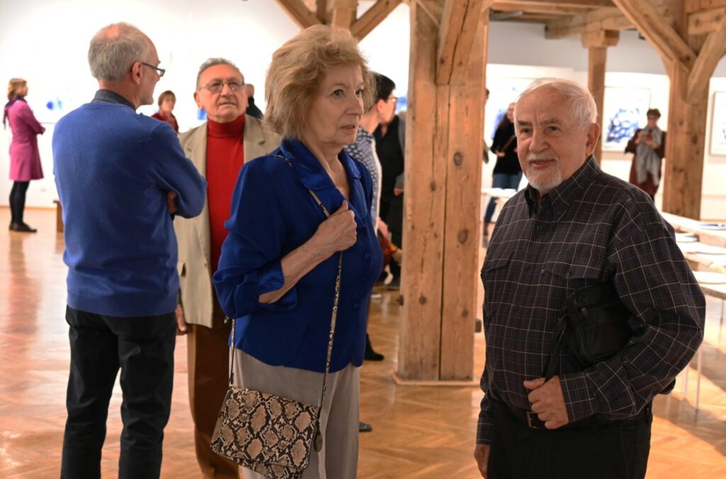 Uczestnicy wernisażu podczas rozmowy w przestrzeni wystawowej; na pierwszym planie widoczna jest kobieta w niebieskiej bluzce oraz mężczyzna, a w tle znajdują się inni goście i elementy ekspozycji (kliknięcie powiększy zdjęcie).