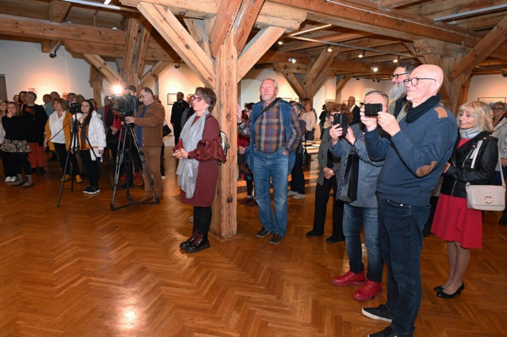 Goście wernisażu zgromadzeni w sali wystawowej; część uczestników fotografuje i nagrywa wydarzenie, a w tle widoczne są drewniane elementy konstrukcji budynku oraz ekspozycja (kliknięcie powiększy zdjęcie).