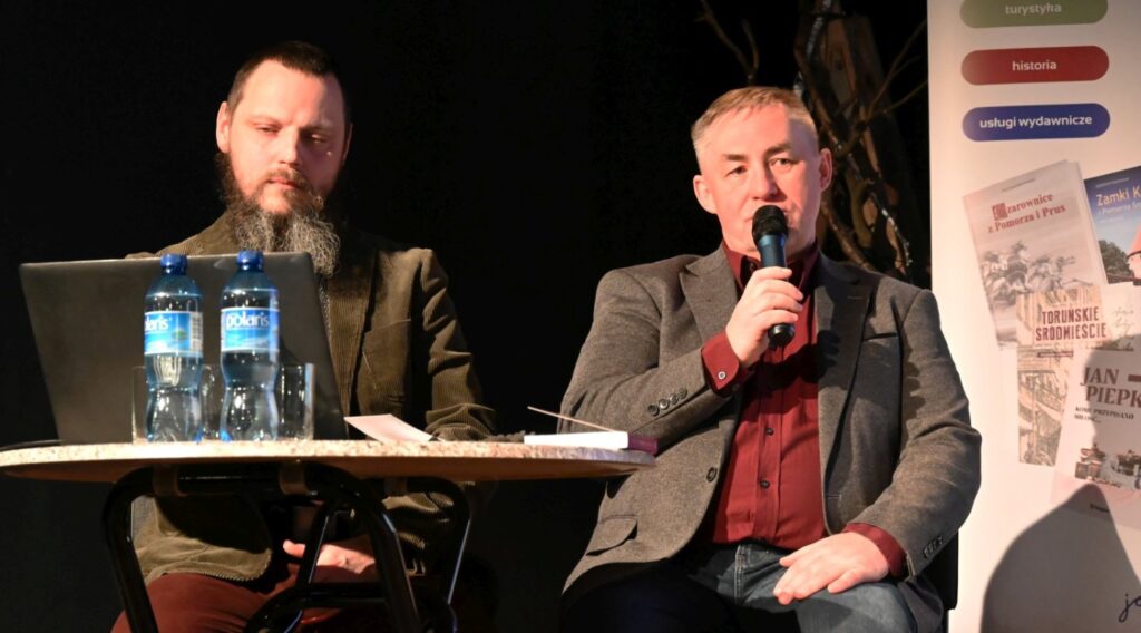 Dwóch mężczyzn siedzi przy stoliku podczas spotkania autorskiego, jeden z nich mówi do mikrofonu (kliknięcie powiększy zdjęcie)