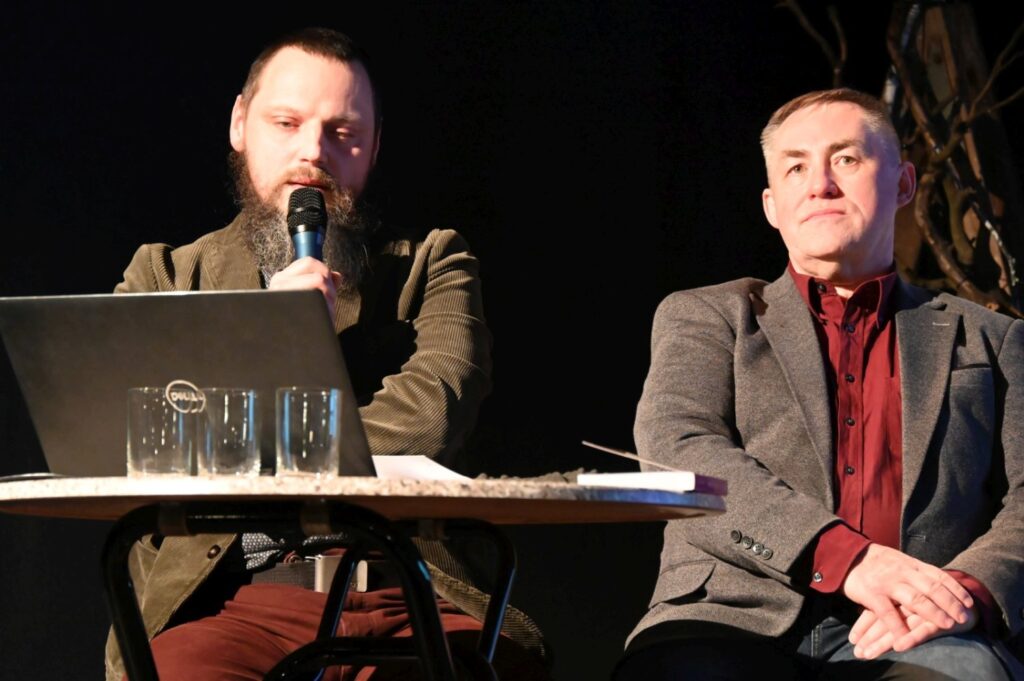 Dwóch uczestników panelu dyskusyjnego siedzi przy stoliku na scenie podczas promocji książki (kliknięcie powiększy zdjęcie).