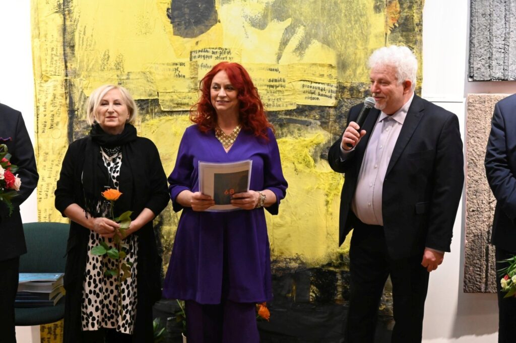 Fotografia z otwarcia wystawy; w centrum kadru stoją przedstawicielka artystów oraz osoby prowadzące wydarzenie, a w tle widoczna jest wielkoformatowa praca malarska w żółtozielonej tonacji; uczestnicy wernisażu uczestniczą w oficjalnej części wydarzenia (kliknięcie powiększy zdjęcie).