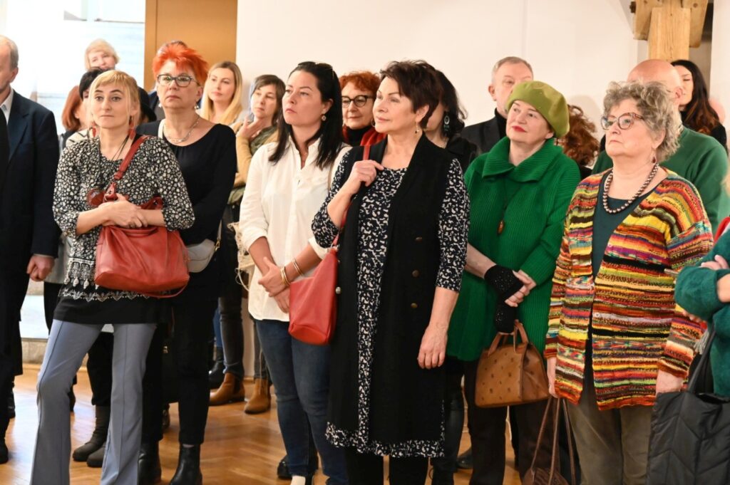 Grupa uczestników zgromadzona podczas wernisażu; publiczność słucha części oficjalnej wydarzenia, stojąc w przestrzeni galerii; w tle widoczne są fragmenty ekspozycji oraz charakterystyczne drewniane elementy konstrukcyjne muzealnego wnętrza (kliknięcie powiększy zdjęcie).
