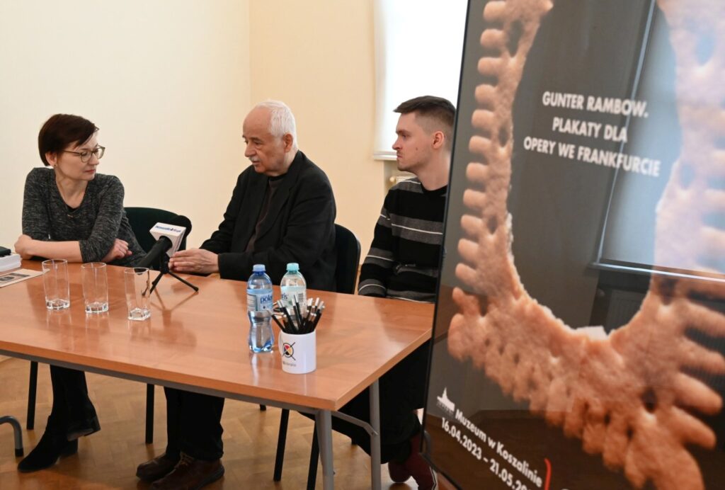 Trzy osoby siedzą przy stole podczas konferencji prasowej obok mikrofony i woda a w tle plakat wystawy (kliknięcie powiększy zdjęcie).