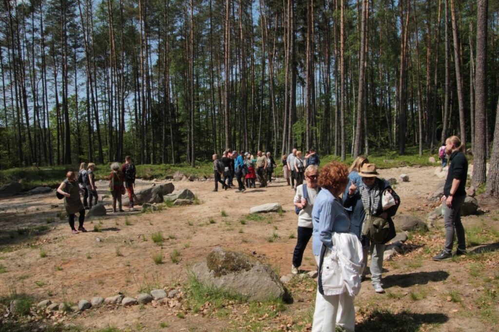 Grupa osób uczestniczących w wycieczce stoi na polanie w lesie wśród wysokich drzew i głazów słuchając przewodnika (kliknięcie powiększy zdjęcie).