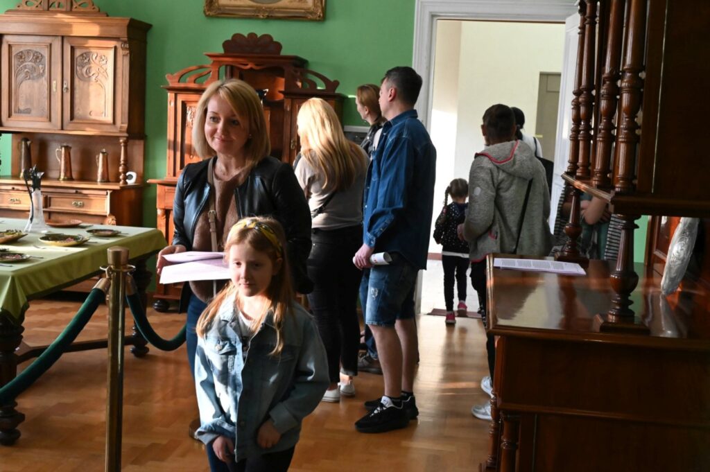 Kobieta z dzieckiem stoi w zabytkowym wnętrzu muzeum a w tle inni zwiedzający oglądają ekspozycję (kliknięcie powiększy zdjęcie).