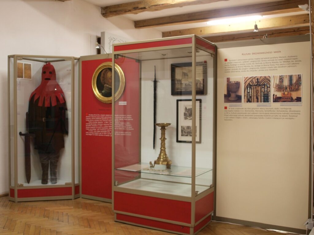 Część wystawy z gablotami prezentującymi historyczne stroje, przedmioty sakralne i elementy wyposażenia; obok znajdują się plansze z fotografiami i informacjami historycznymi (kliknięcie powiększy zdjęcie).