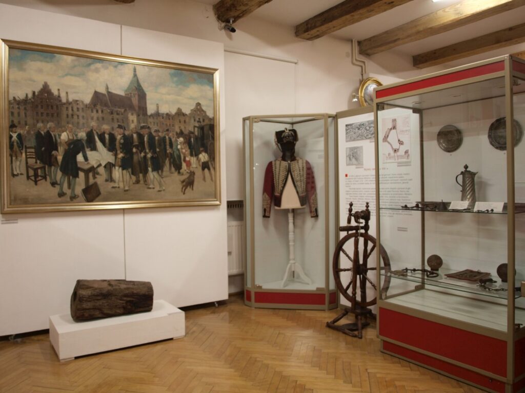 Fragment wystawy z dużym historycznym obrazem przedstawiającym scenę miejską; obok znajdują się manekin w dawnym stroju, kołowrotek i inne eksponaty w gablocie (kliknięcie powiększy zdjęcie).