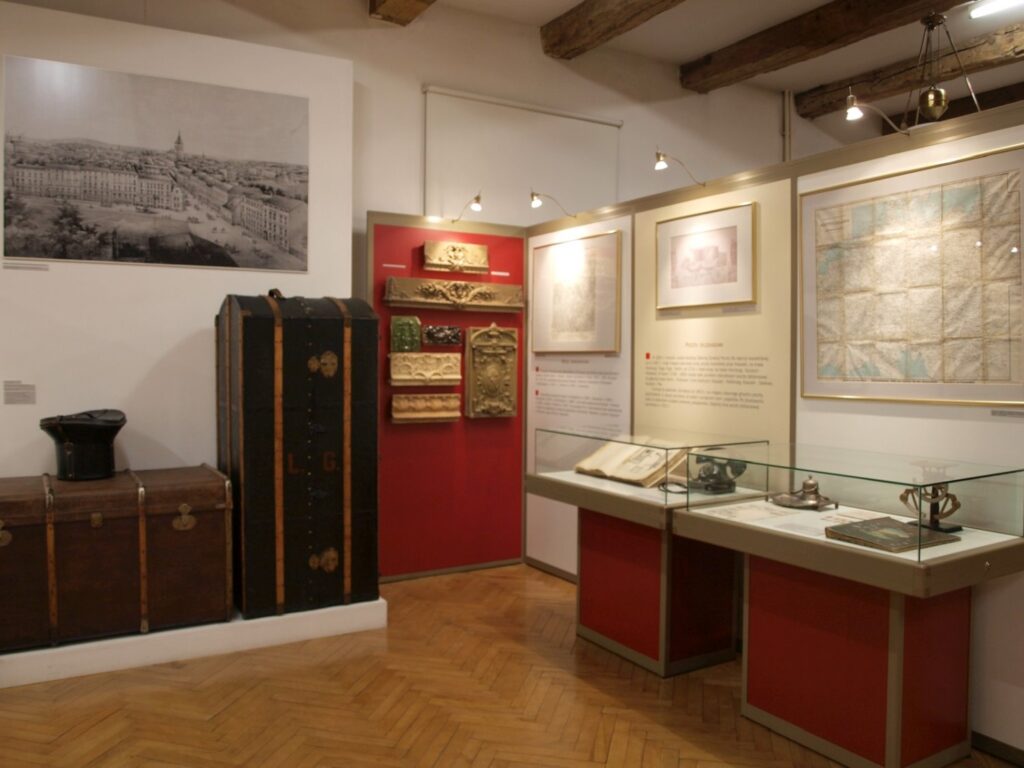 Fragment ekspozycji z zabytkowymi skrzyniami, dokumentami, mapami i przedmiotami umieszczonymi w gablotach; na ścianie widoczna jest duża historyczna fotografia miasta (kliknięcie powiększy zdjęcie).