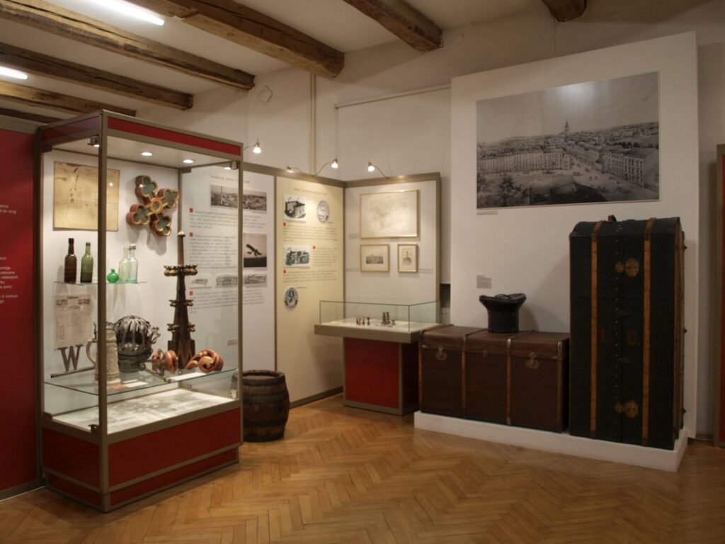 Sala wystawowa z gablotami prezentującymi dawne przedmioty, naczynia, dokumenty i pamiątki; na ścianach znajdują się fotografie, ilustracje i teksty dotyczące historii miasta (kliknięcie powiększy zdjęcie).