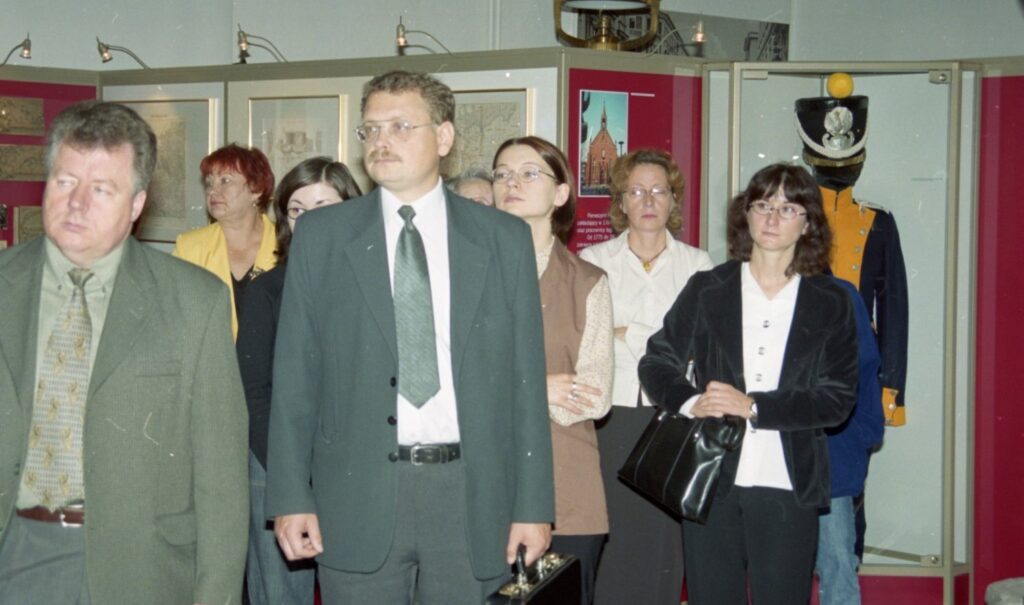 Uczestnicy wydarzenia zwiedzają wystawę; w centrum znajduje się grupa gości, a w tle widoczne są gabloty z eksponatami i elementy ekspozycji (kliknięcie powiększy zdjęcie).