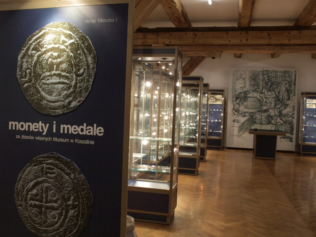 Plakat wystawy „Monety i medale”, prezentujący wizerunki dwóch monet; w tle rząd podświetlonych gablot z numizmatami oraz wielkoformatowa grafika o tematyce historycznej (kliknięcie powiększy zdjęcie).