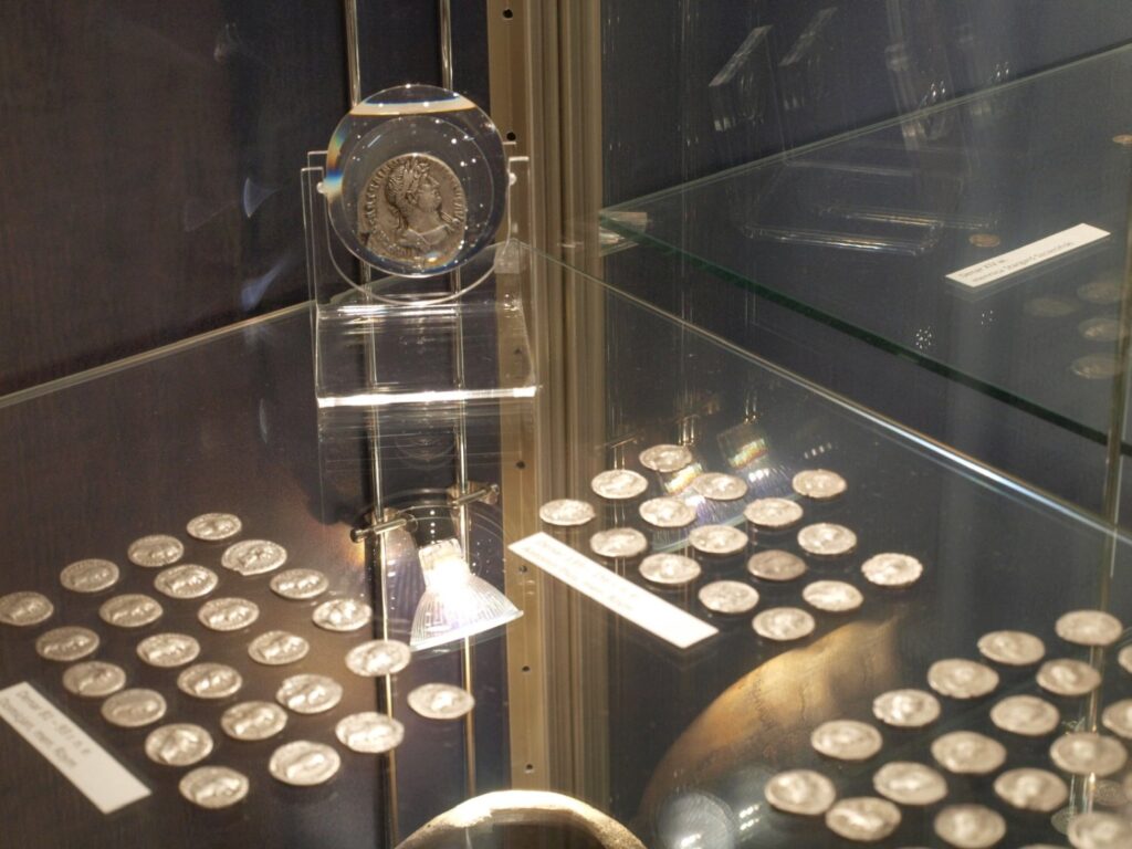 Fragment gabloty z pojedynczą monetą umieszczoną na przezroczystym postumencie z możliwością oglądania jej przez lupę; poniżej i w głębi ekspozycji widoczne są liczne drobne monety rozmieszczone w uporządkowanych grupach oraz tabliczki z opisami (kliknięcie powiększy zdjęcie).