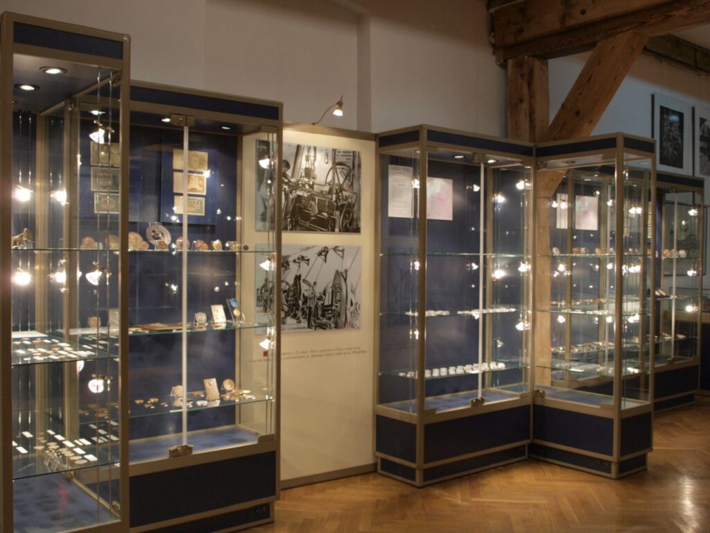 Fragment wystawy numizmatycznej z wysokimi, podświetlonymi gablotami; wewnątrz widoczne są monety, medale, dokumenty i ilustracje związane z historią pieniądza; ekspozycję uzupełniają ciemne plansze i grafiki umieszczone na ścianach (kliknięcie powiększy zdjęcie).
