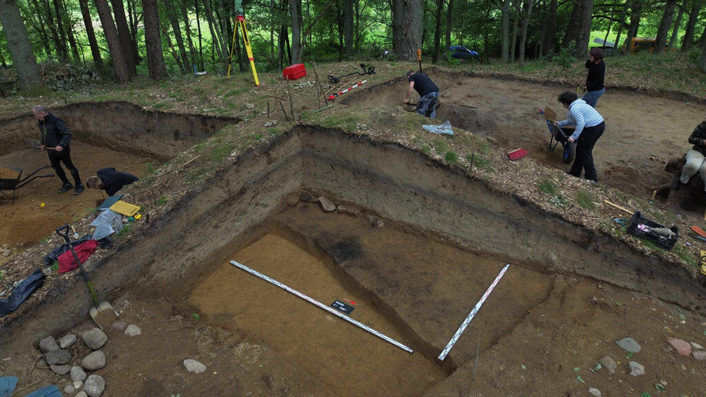 Archeolodzy pracują w wykopie w lesie, odsłaniając warstwy ziemi i dokumentując znaleziska (kliknięcie powiększy zdjęcie).