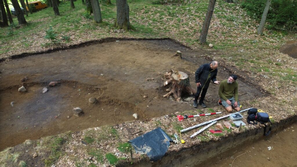Archeolodzy mierzą i dokumentują znaleziska w wykopie, korzystając z narzędzi pomiarowych (kliknięcie powiększy zdjęcie).