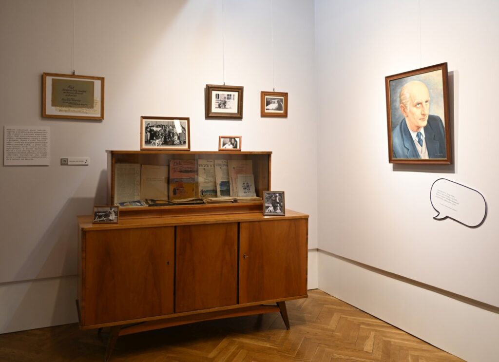 Fragment wystawy z drewnianą szafką i oprawionymi materiałami archiwalnymi na ścianach; w gablocie widoczne są książki i dokumenty, nad nią zawieszono fotografie, a po prawej stronie znajduje się portret mężczyzny uzupełniony o graficzny dymek z wypowiedzią; całość tworzy aranżację przypominającą dawne wnętrze mieszkalne lub gabinet (kliknięcie powiększy zdjęcie).
