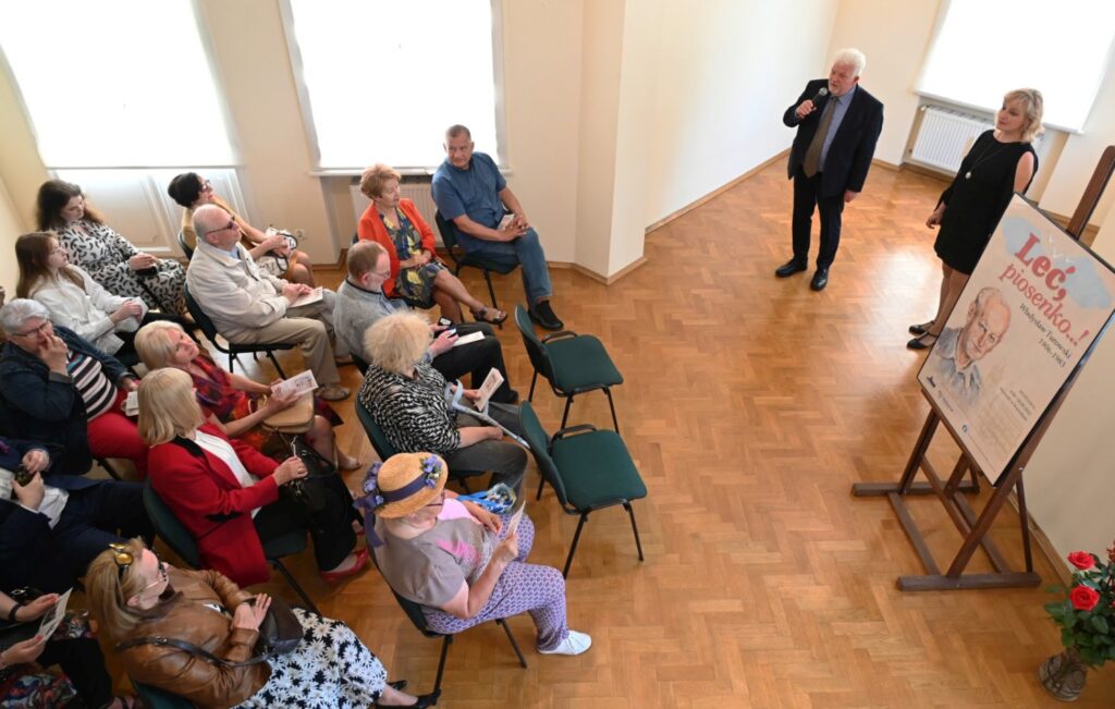 Otwarcie wystawy w sali wypełnionej publicznością; po lewej stronie uczestnicy siedzą na krzesłach ustawionych w kilku rzędach i słuchają przemawiającego mężczyzny stojącego przy ścianie; obok znajduje się plansza z plakatem wystawy (kliknięcie powiększy zdjęcie).